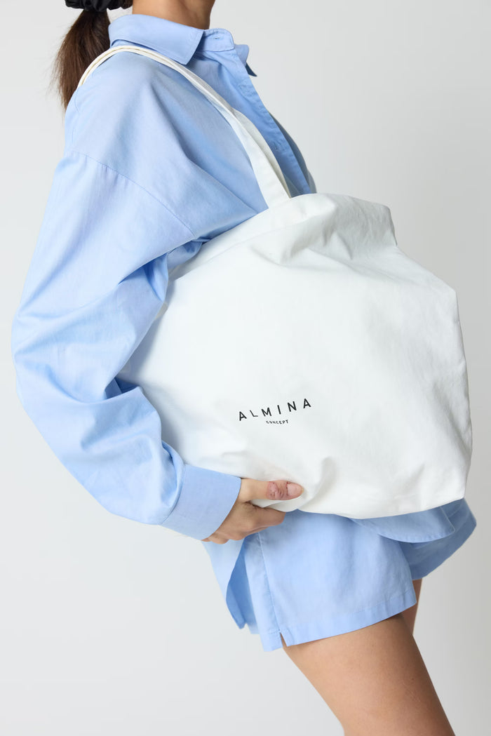 Person wearing a light blue shirt and shorts holding a white tote bag with 'ALMINA' branding.