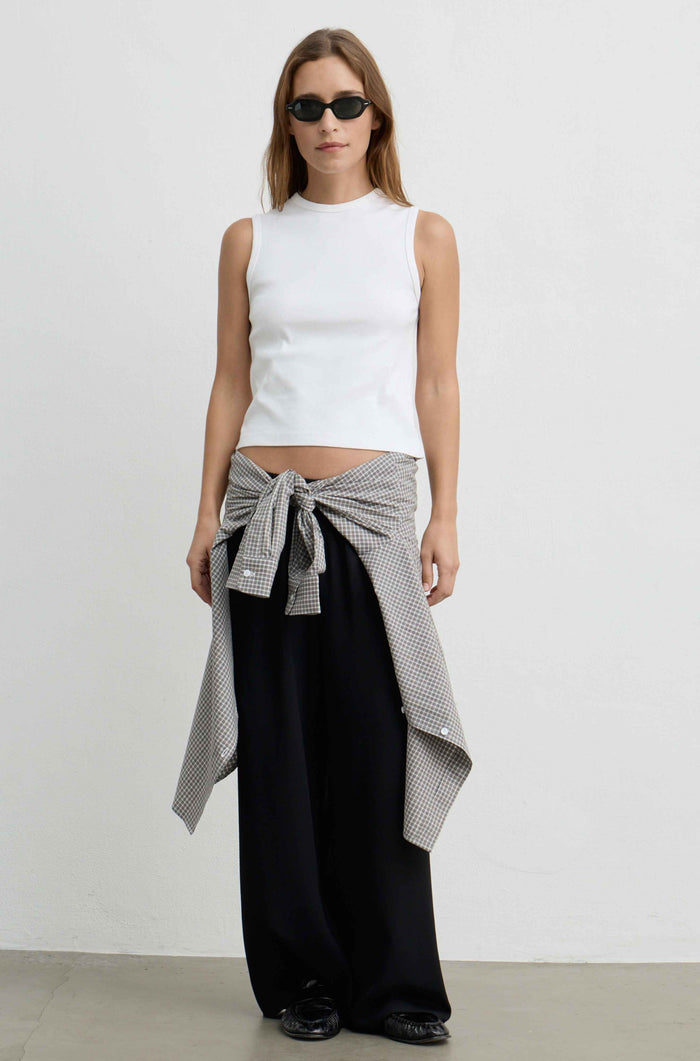 A woman stands against a plain light gray wall wearing a white sleeveless mock-neck top, loose black pants, black sunglasses, and a gray-and-white patterned shirt tied around her waist.