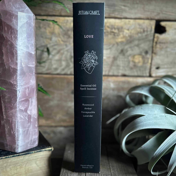 A box of 'Love Spell Incense Sticks' by Ritualcraft, featuring an illustration of a flower superimposed on a heart, placed on a wooden surface with a rose quartz crystal and and air plant.