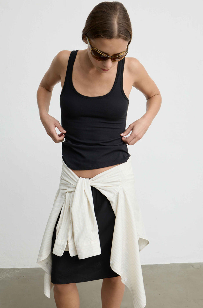 A woman wearing a black tank top and black skirt, with a cream-colored knit wrap tied around her waist, set against a plain light gray studio background.