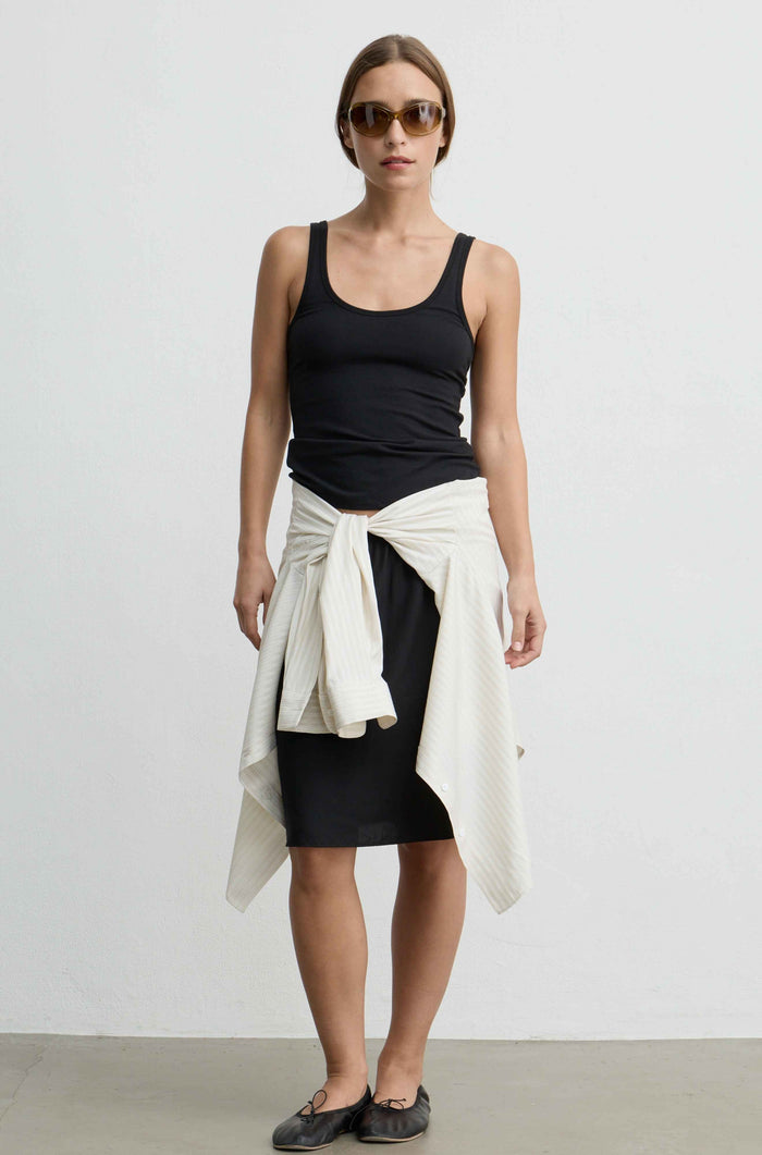 A woman modeling a black tank top, black skirt, and a cream-colored knit sweater tied around her waist, paired with dark flat shoes and sunglasses, against a plain white studio background.
