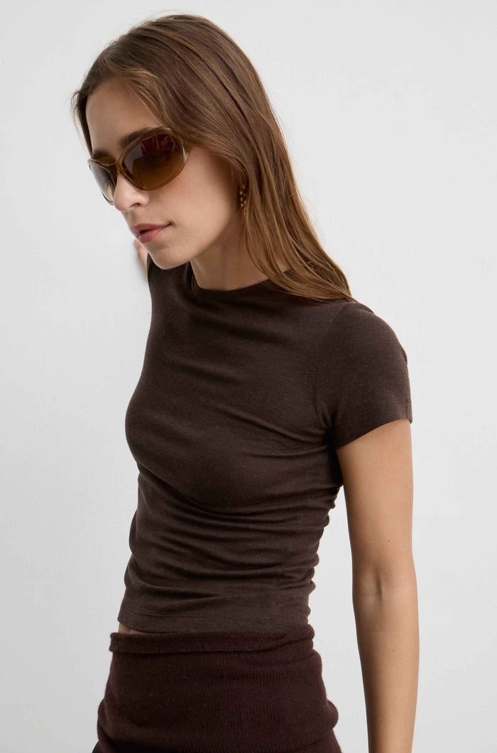 A woman wearing a fitted short-sleeve ribbed knit top in dark brown, paired with matching dark brown bottoms and gold-framed sunglasses, photographed against a plain light gray studio background.