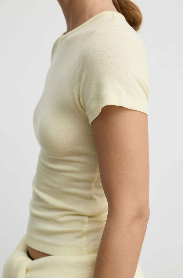A close-up side view of a person wearing a pale butter yellow short-sleeve ribbed knit top against a plain light background.