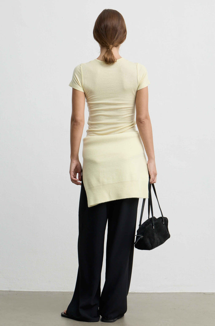 A woman shown from the back wearing a pale yellow short-sleeve knit top and black wide-leg trousers, holding a small black handbag against a plain light gray studio background.