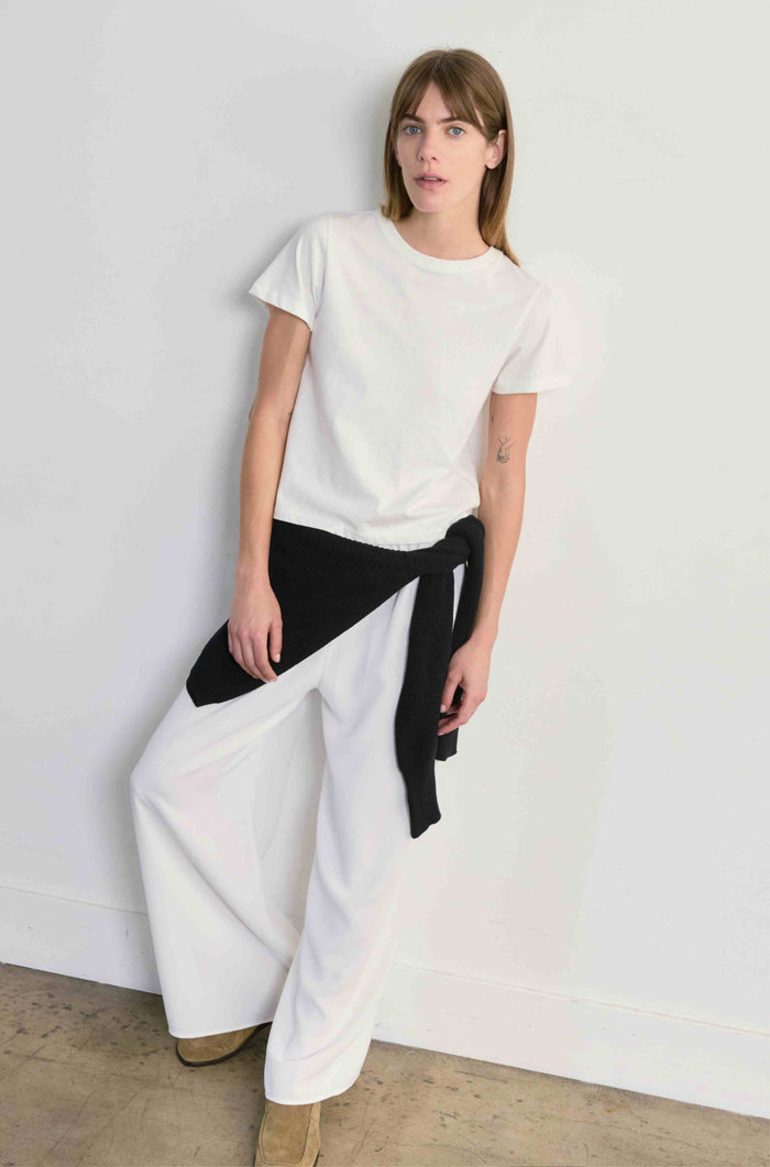 Woman wearing a white top and wide-leg pants with a black belt against a plain background